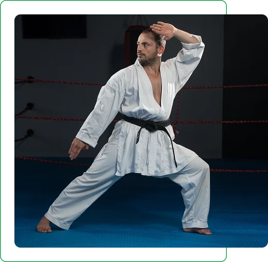 Un homme en gi de karaté blanc et ceinture noire adopte une posture d'arts martiaux sur un tapis bleu, encadré par des cordes rouges, avec un regard intense et concentré.